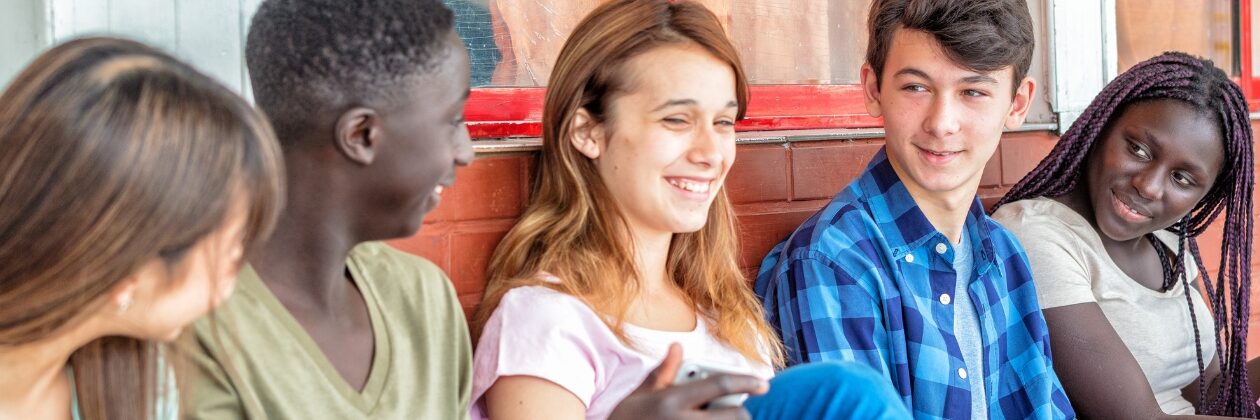 Photo of 5 teenagers, a mixture of genders and skin colours, smiling and chatting to one another.