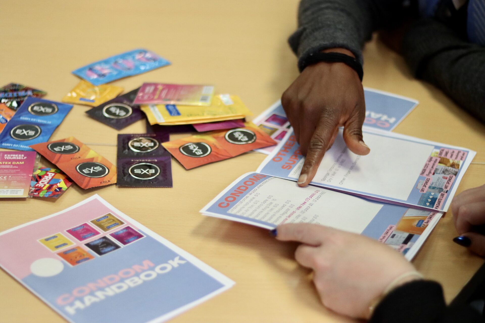 Close up of table and two pair of hands pointing at a booklet with condom prices. On the table are various types of condoms.